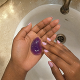 Close-up of purple jelly cleanser texture in hands above a bathroom sink.