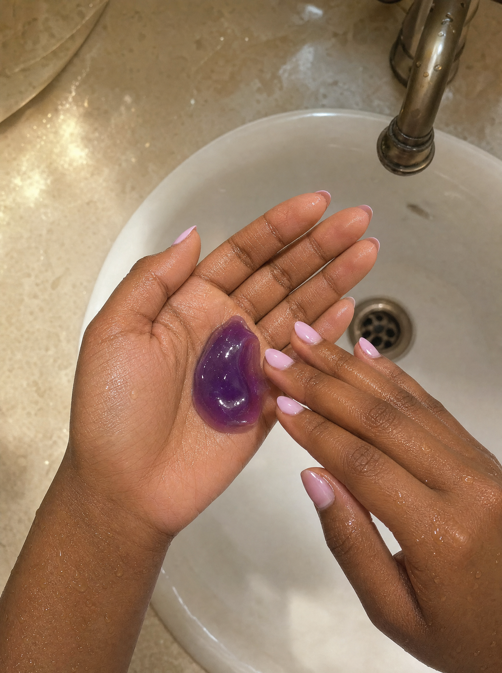 Close-up of purple jelly cleanser texture in hands above a bathroom sink.